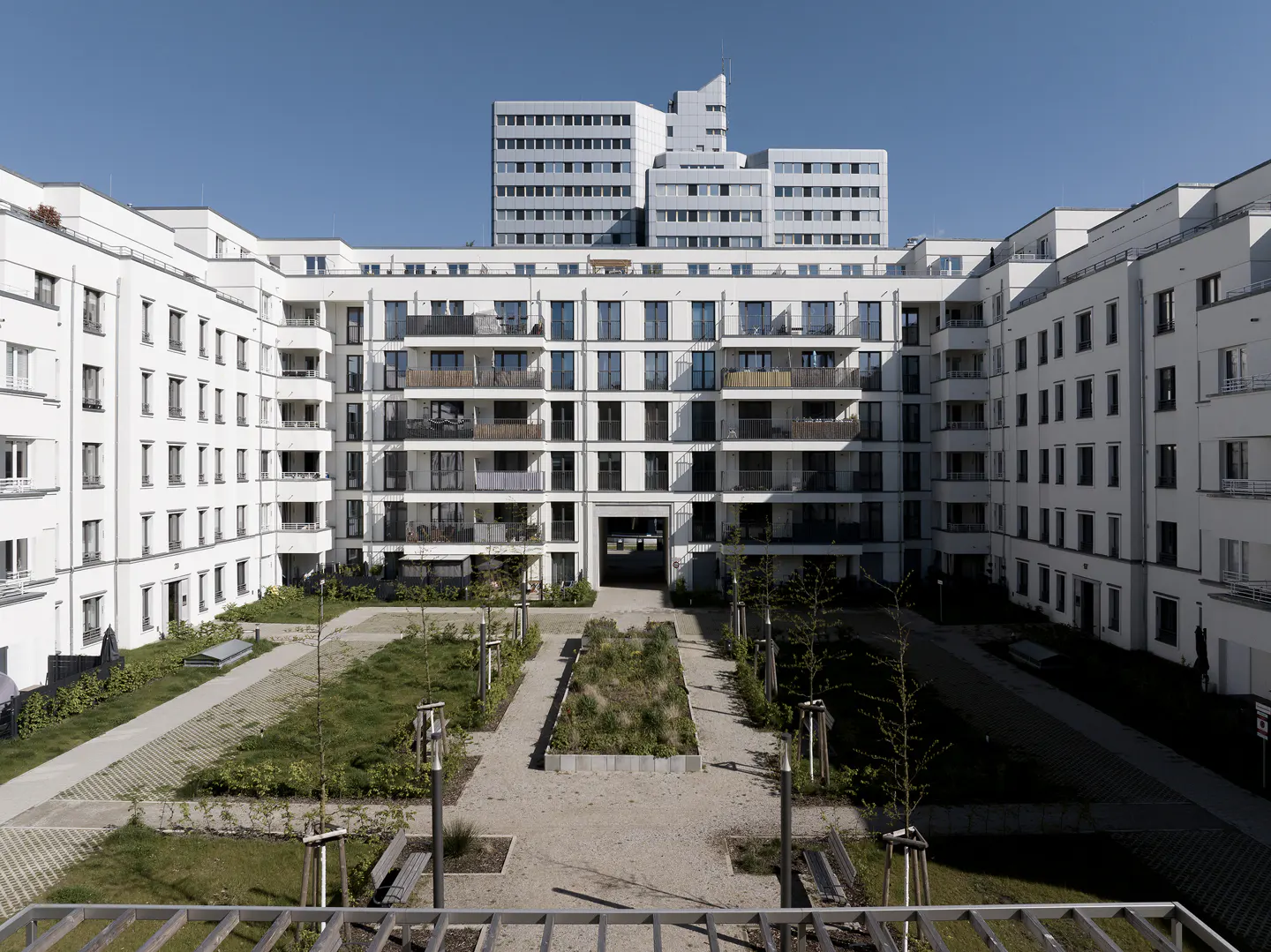 View of a modern white apartment complex with balconies surrounding a courtyard with grass and trees under a clear blue sky.