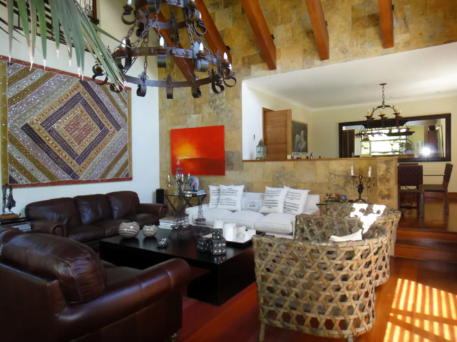 Living room with brown leather sofas, white sofa, woven chairs, black coffee table, and stone walls. A large chandelier hangs from the ceiling.