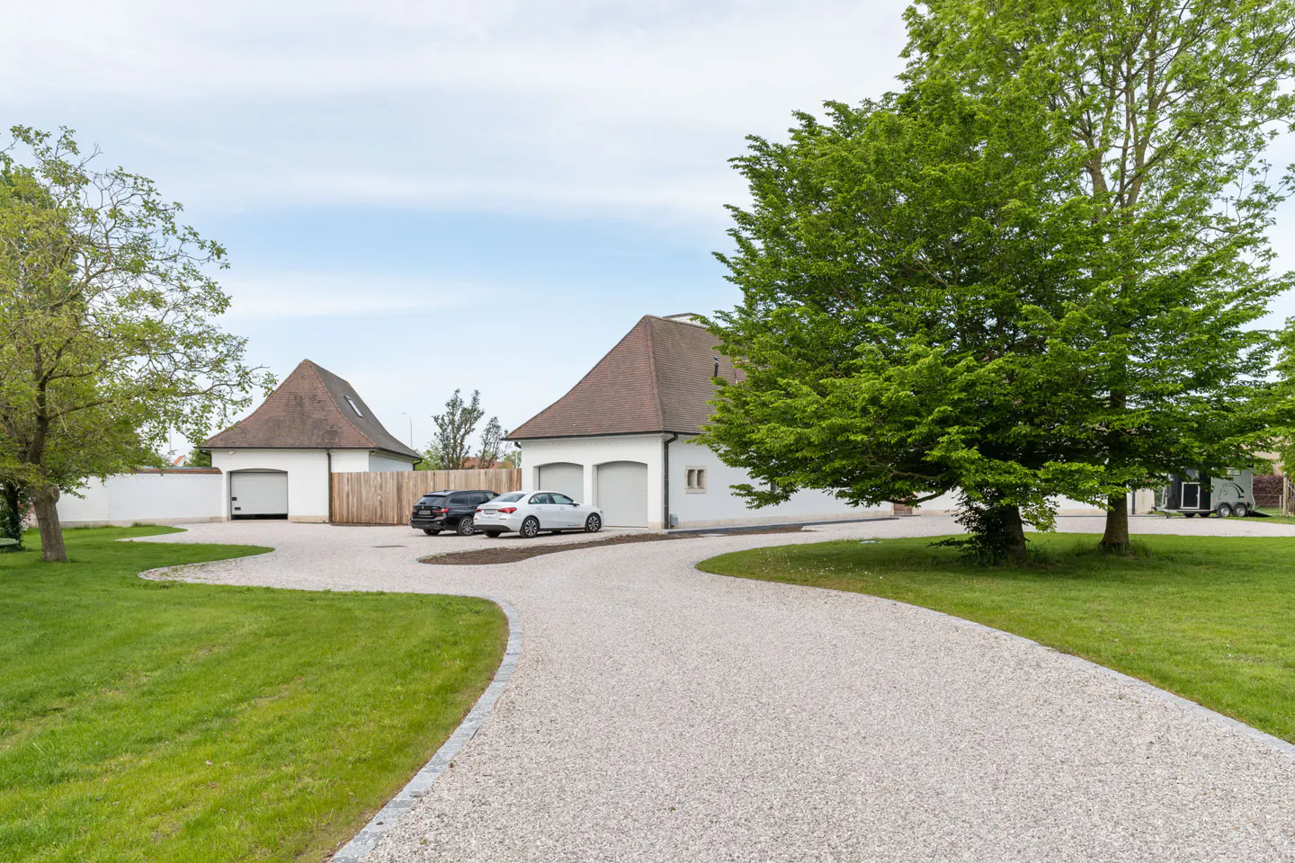 Exterior view of a white house with brown roof, a gravel driveway, green lawn, and two cars parked in front of the garage.