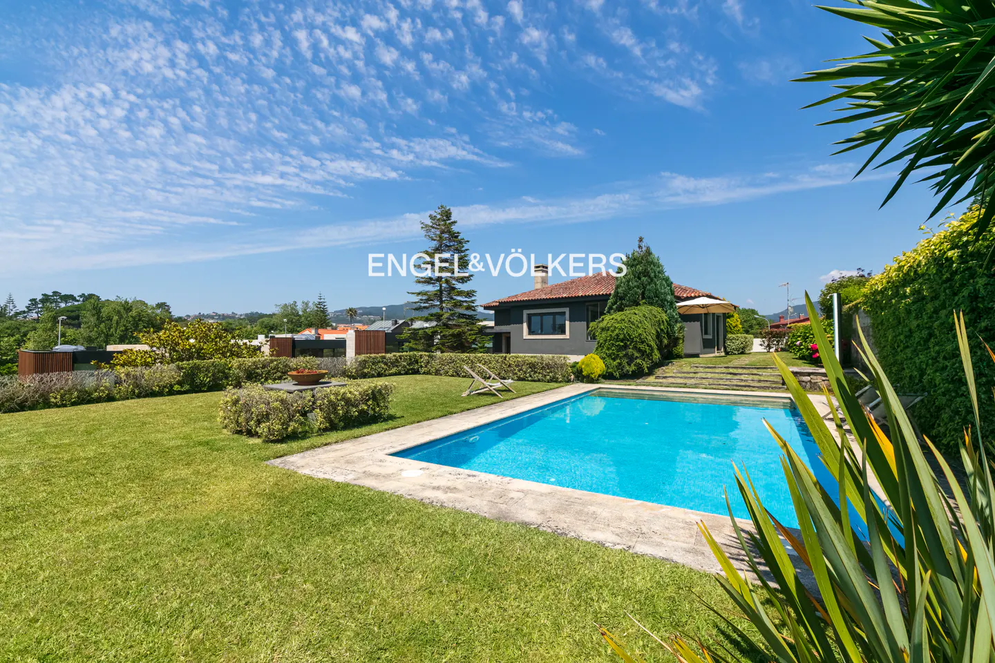 A backyard with a blue pool, green grass, and a gray house with a red roof under a blue sky.