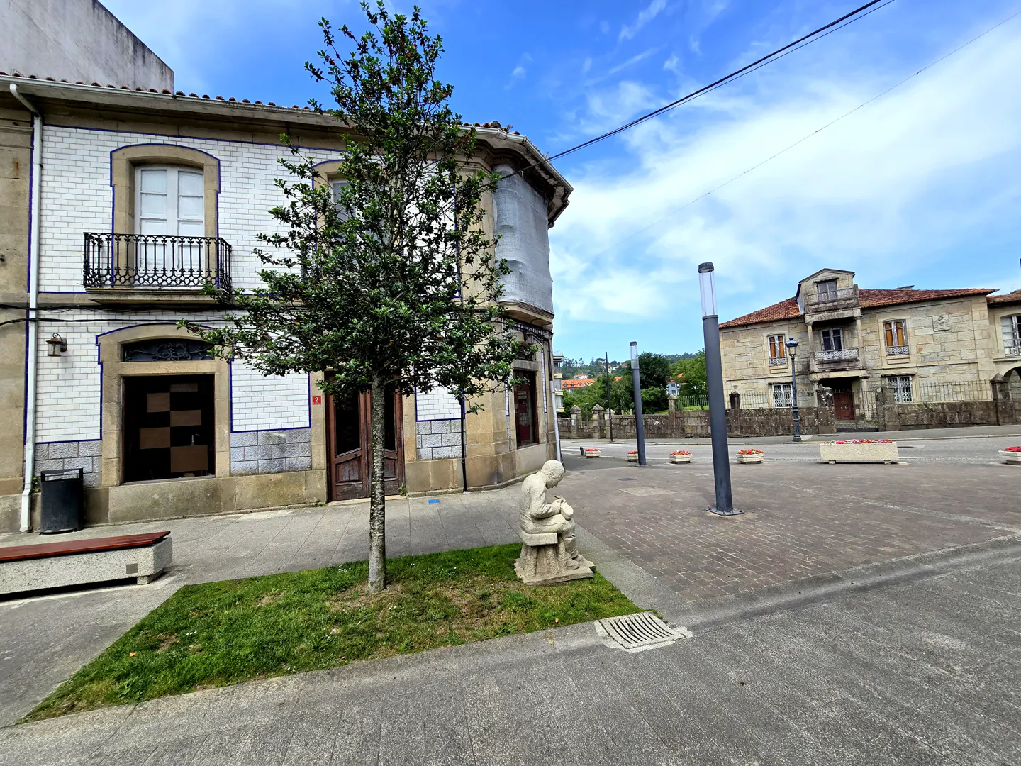Exterior view of a two-story building with white tiled walls, a tree, and a statue of a person sitting on a stool.