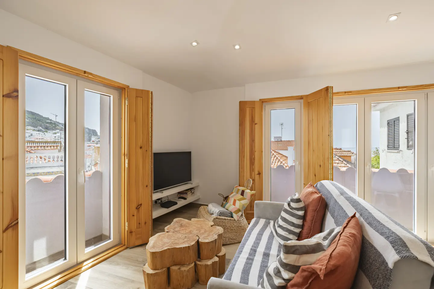 Bright living room with wood-framed windows, a gray sofa with striped pillows, and a unique wooden coffee table. A TV sits on a white stand.