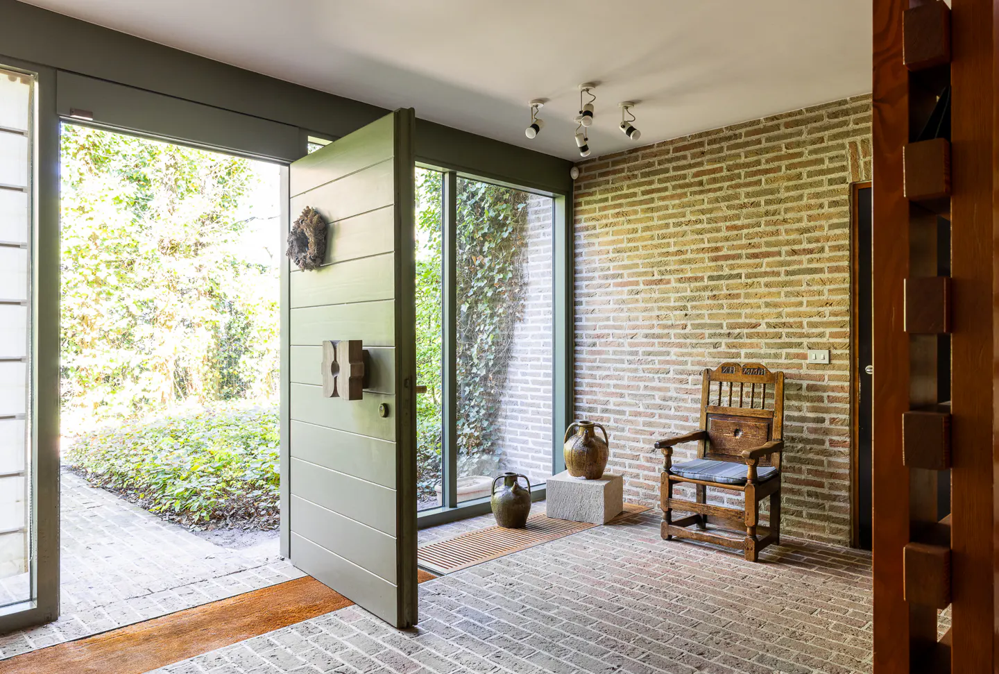 A modern home's entryway with an open sage door, brick floor and wall, and a wooden chair.