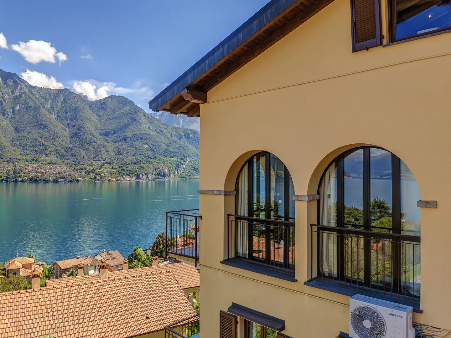 Lake view from a yellow building with arched windows and black railings. Mountains and blue water fill the background.