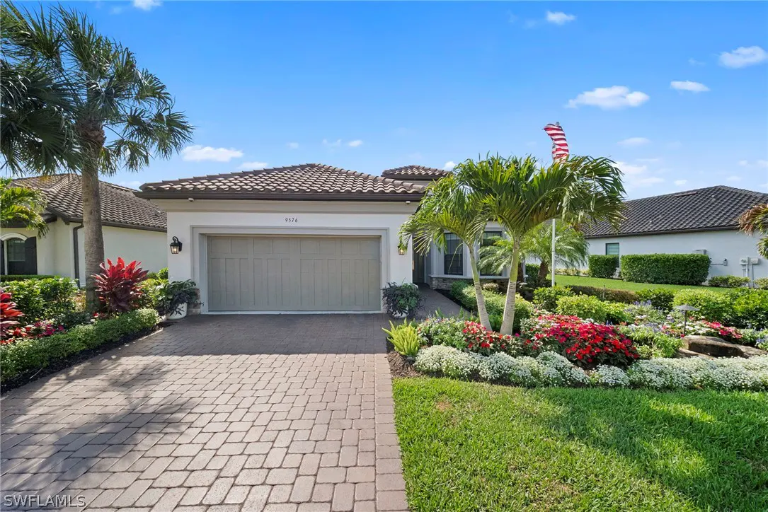 Exterior of a one-story white house with a brown tile roof, a two-car garage, and a brick driveway. Palm trees and colorful flowers surround the house.