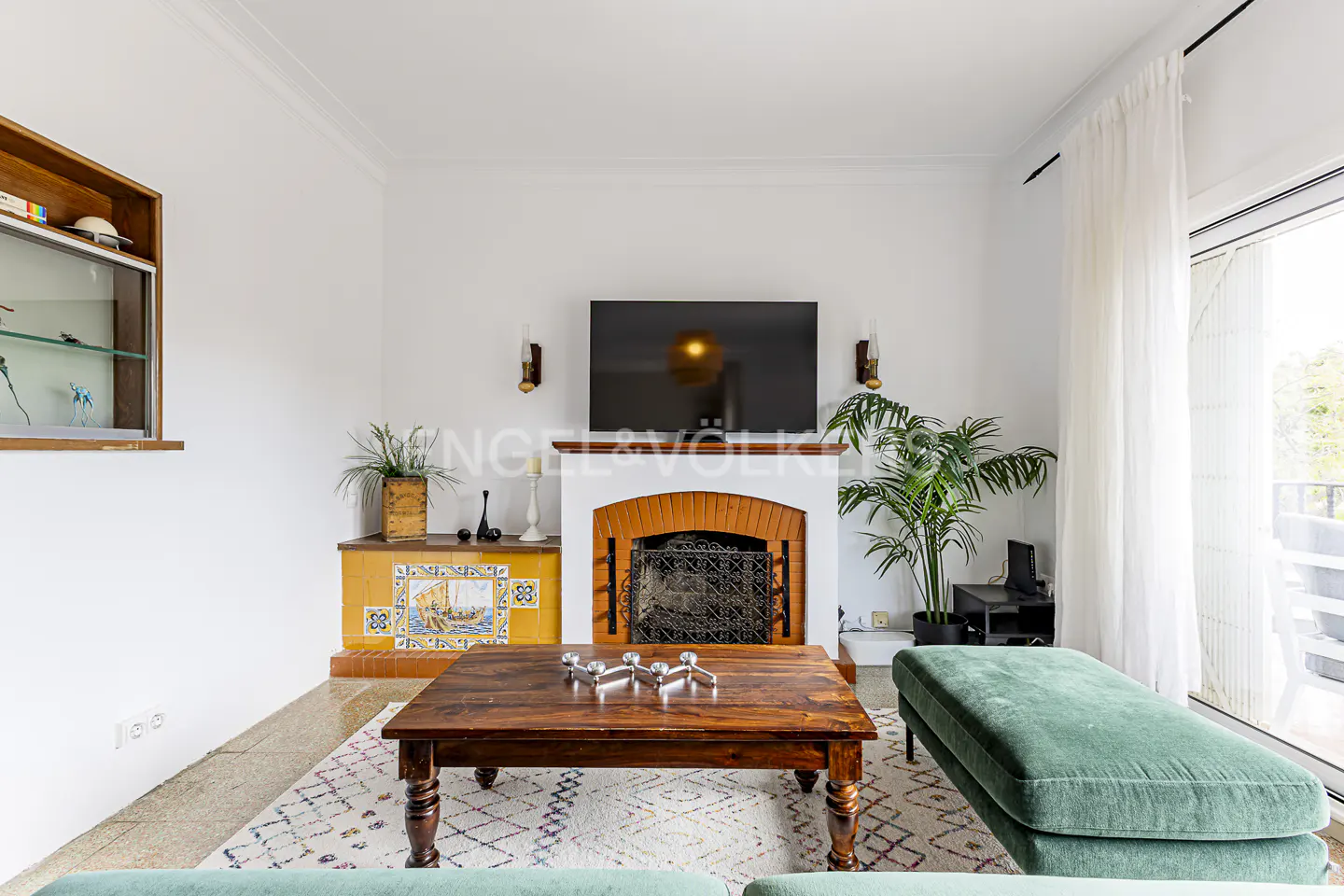 Living room with white walls, a brick fireplace, a dark wood coffee table, and a green ottoman. A TV hangs above the fireplace.