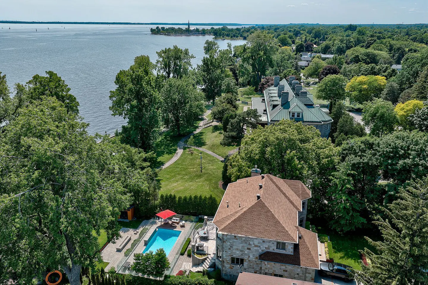 Aerial view of a stone house with a brown roof, a blue pool, and a red umbrella, surrounded by green trees and a lake.