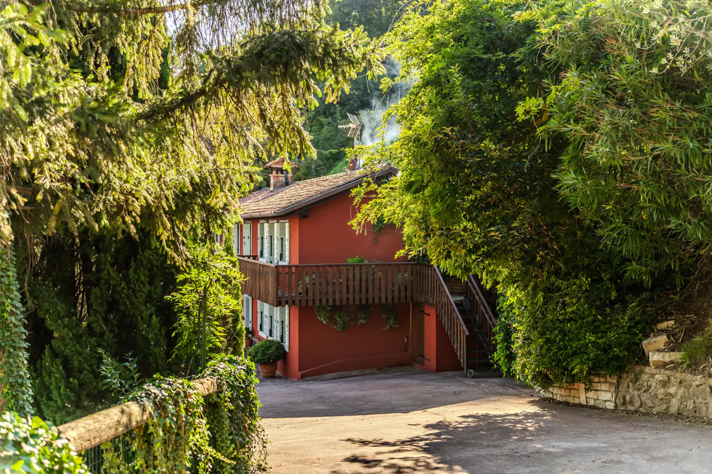Red house with brown roof and white shutters, surrounded by green trees and foliage. A wooden balcony and stairs add to the charm.