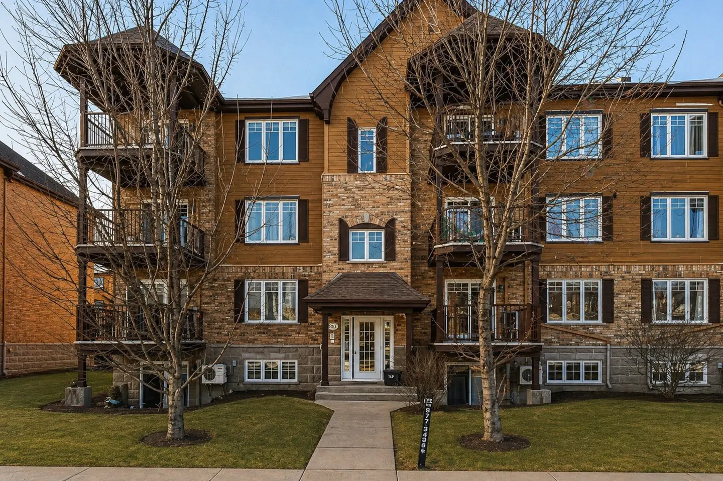 Three-story apartment building with brown wood and brick facade, white windows, and small balconies. Two bare trees frame the building.