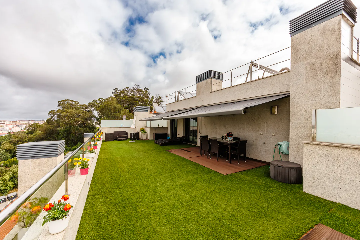 Spacious rooftop terrace with artificial grass, outdoor dining set, and potted flowers overlooking a wooded area.