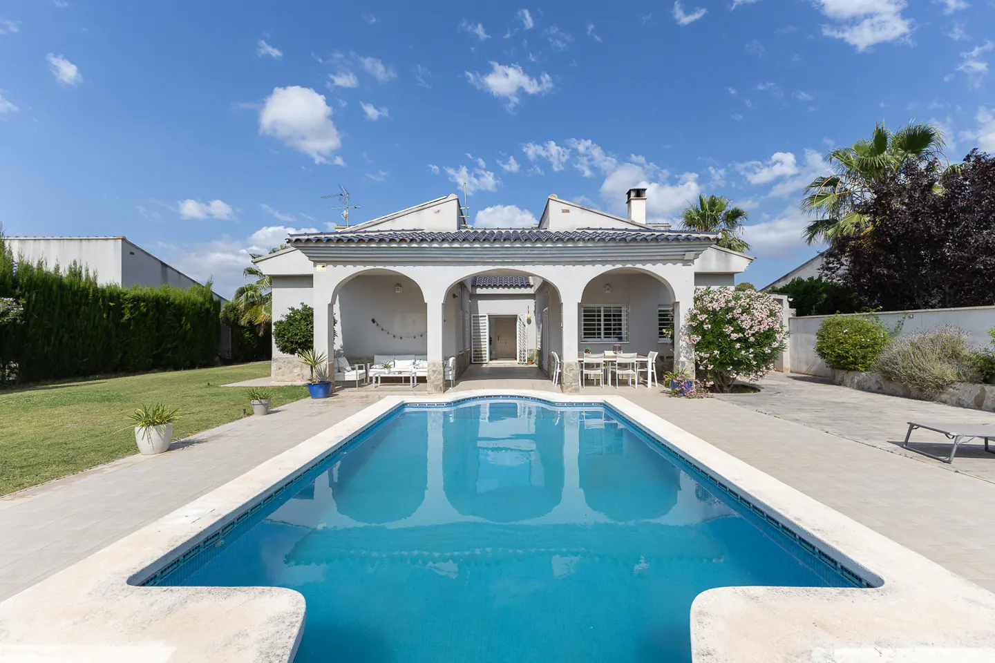Backyard view of a white villa with a blue swimming pool, covered patio, and green lawn under a bright blue sky.