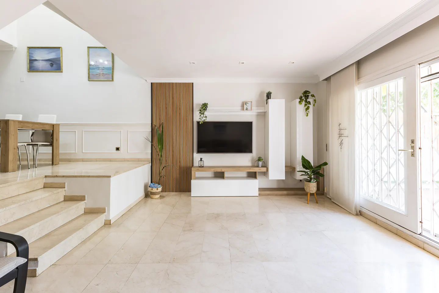 Bright living room with marble floors, white walls, and a wooden accent wall. A TV is mounted above a white cabinet with plants. A dining area is elevated by stairs.