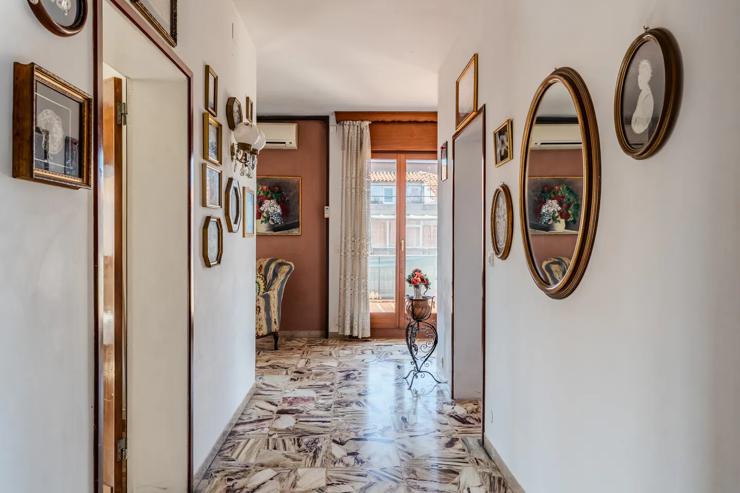 Hallway with marble floors and white walls decorated with framed art and mirrors. A doorway leads to a room with a window and chair.