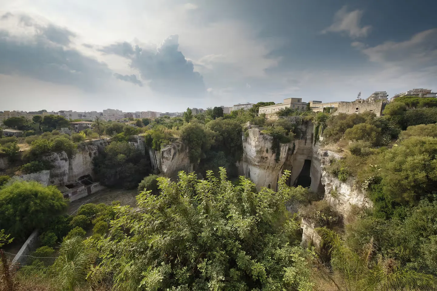 Scenic view of Latomia dei Cappuccini, a quarry in Syracuse, Italy, with lush greenery, white cliffs, and buildings in the background under a cloudy sky.