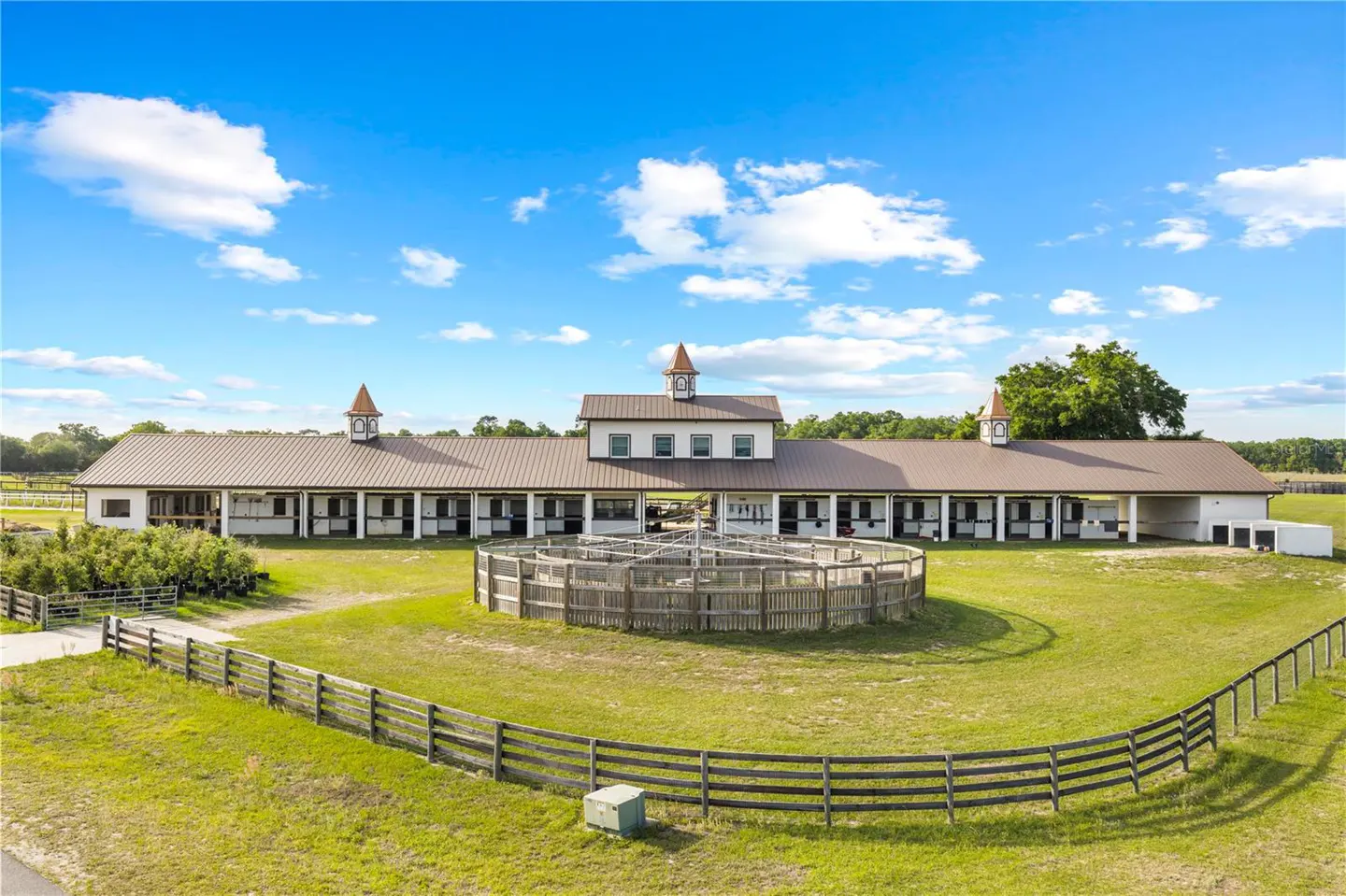 Exterior view of a white horse barn with a brown roof, a round pen, and a wooden fence in a grassy field.