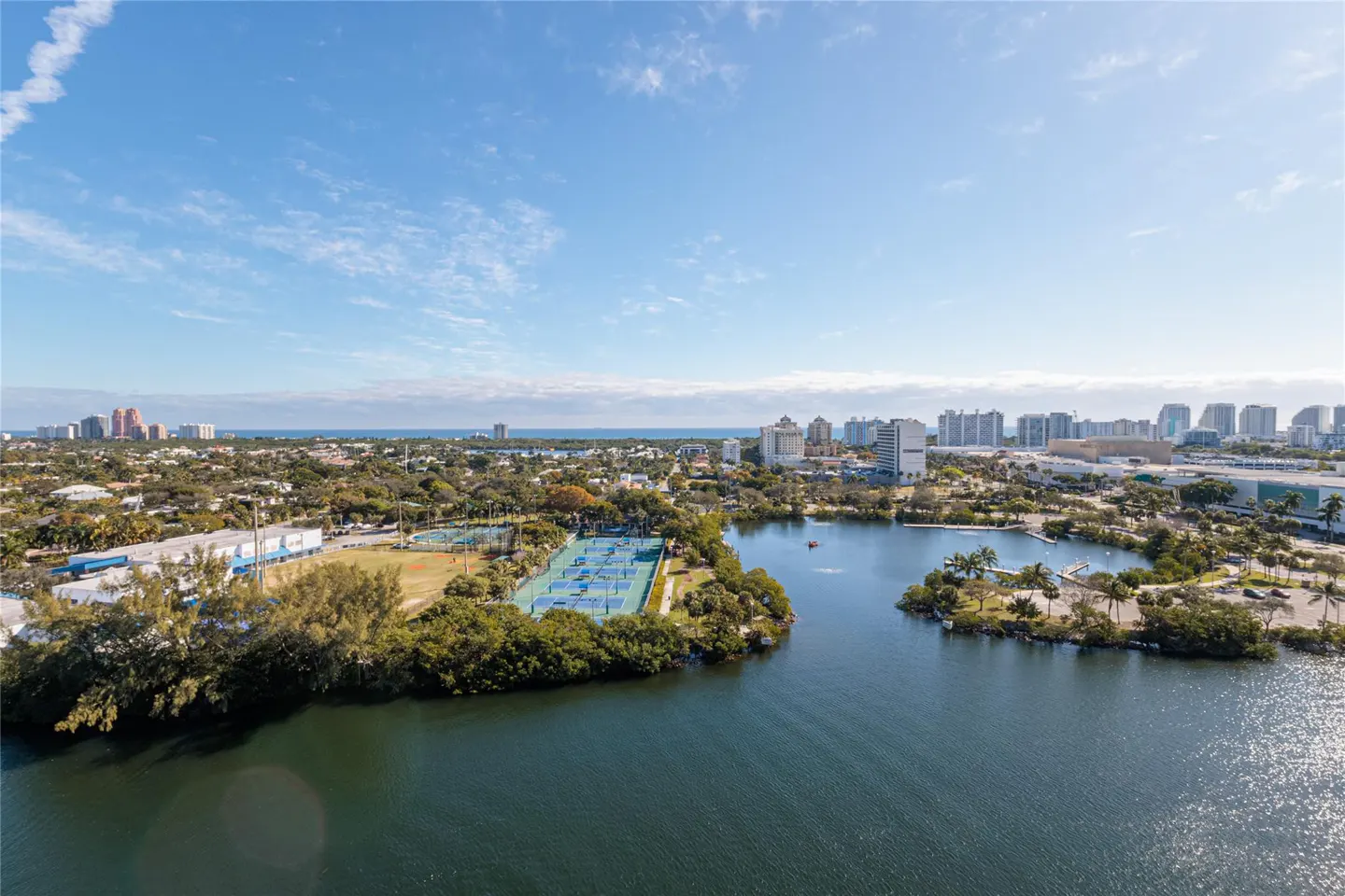 Aerial view of a city with a lake, tennis courts, and buildings under a blue sky with scattered clouds.