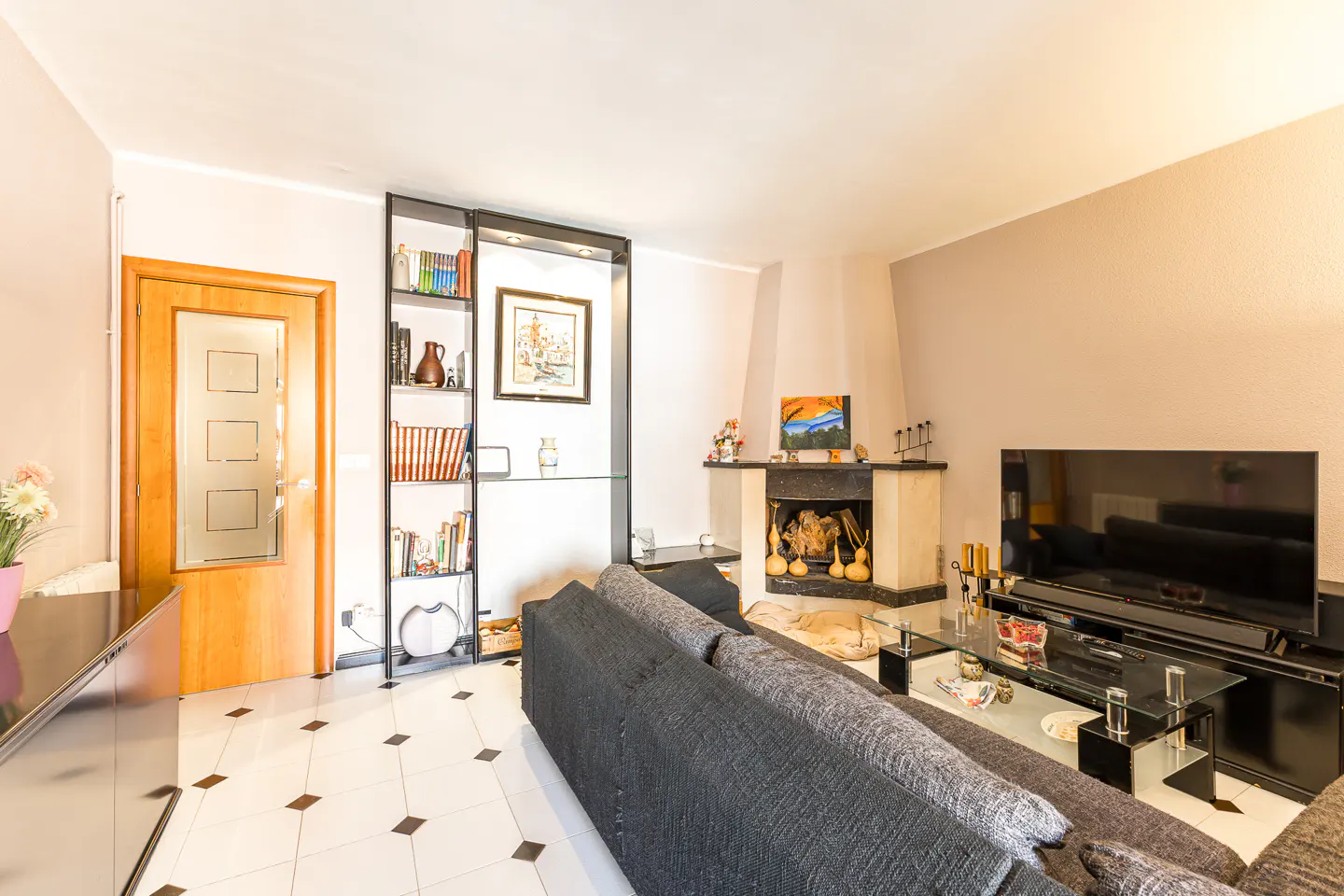 Living room with a gray sofa, fireplace, TV, and bookshelf. The floor is white tile with black accents. A wooden door is on the left.