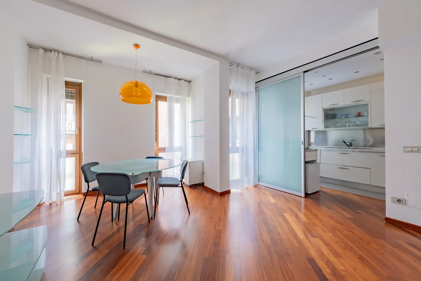 Bright apartment with wood floors, a glass table with four chairs, and a sliding glass door to a white kitchen.
