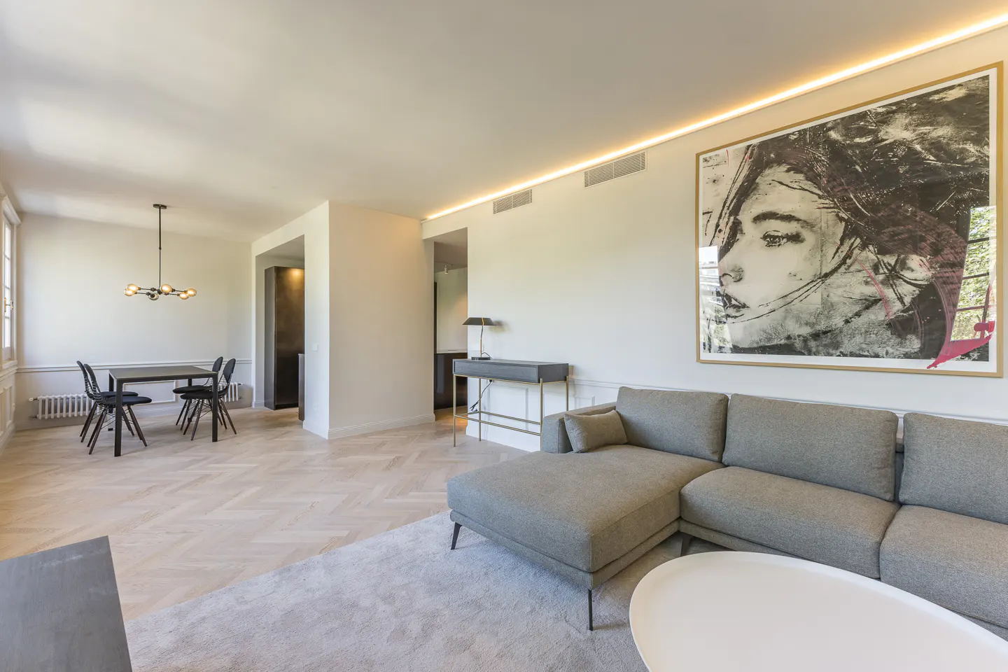 Bright living room with gray sectional sofa, white oval coffee table, and large portrait art. Dining table with black chairs in background.