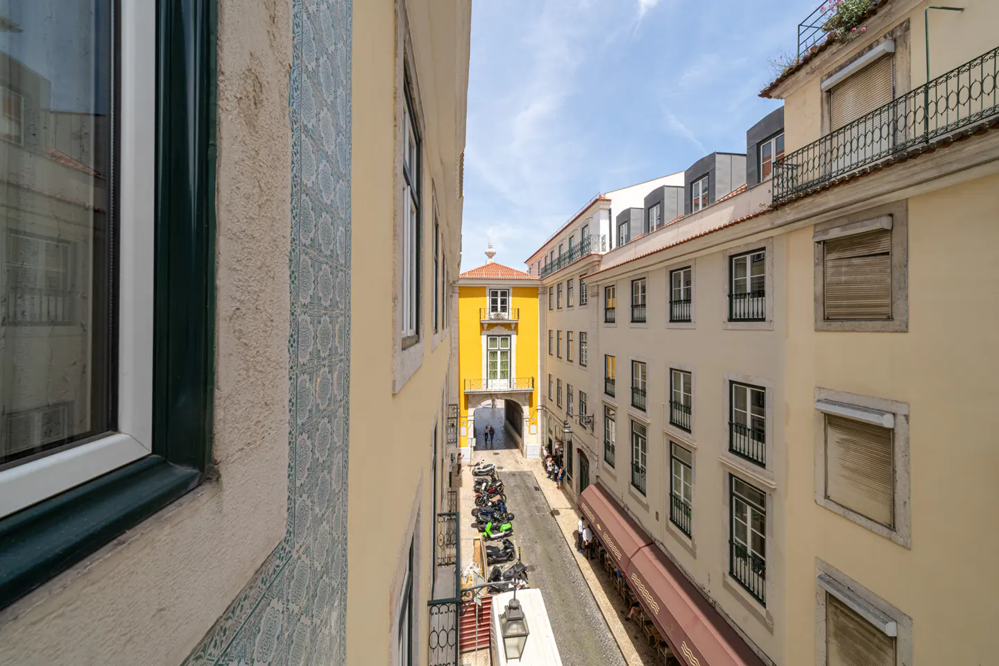 View from a window of a narrow street in Lisbon, Portugal, with colorful buildings and parked scooters.