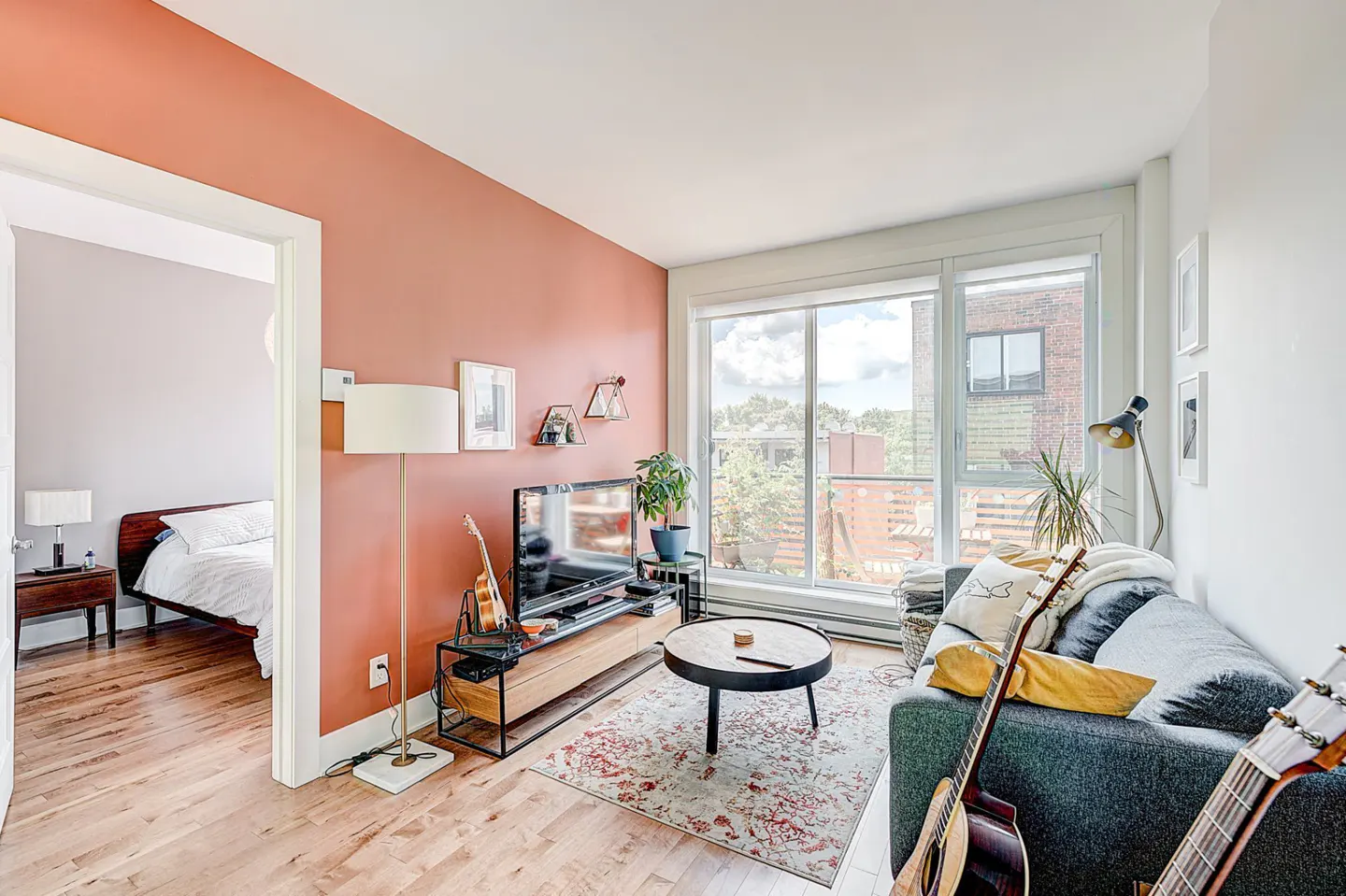 Living room with a gray sofa, guitars, and a TV. A salmon-colored wall adds warmth, while a large window offers a city view. Hardwood floors and a floral rug complete the space.