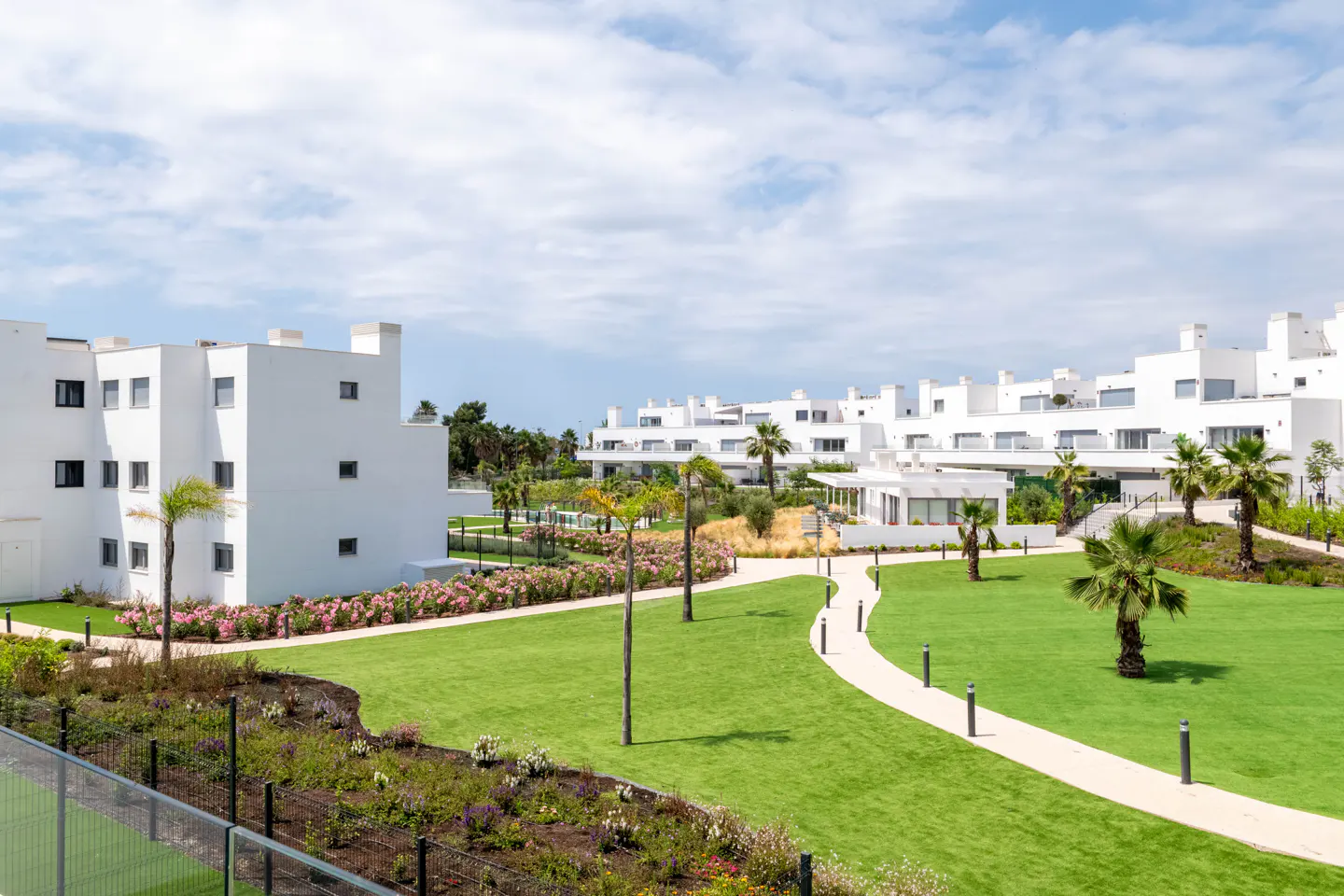 Modern white apartment buildings surrounded by green lawns, palm trees, and landscaped flower beds under a blue sky.
