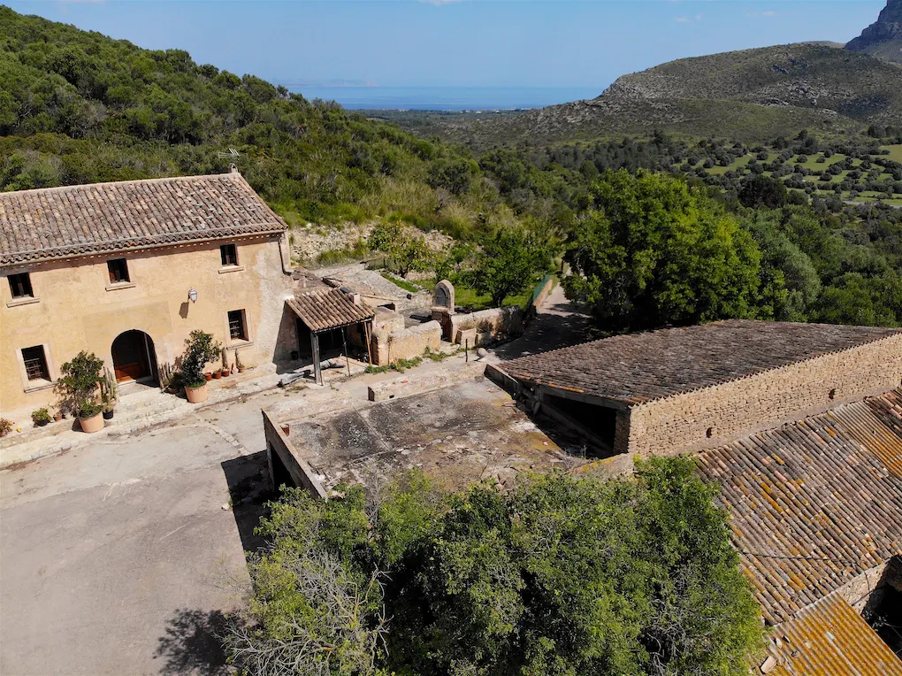 Aerial view of a tan stone house with a tile roof, surrounded by green trees and hills under a blue sky.