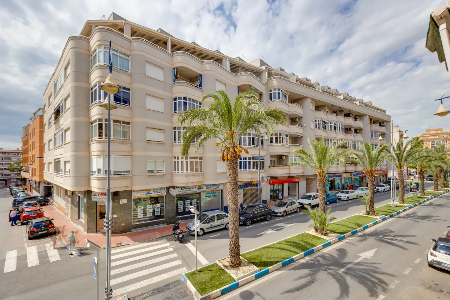 Street view of a multi-story apartment building with palm trees lining the street. Cars are parked along the curb. Pedestrians cross the street.