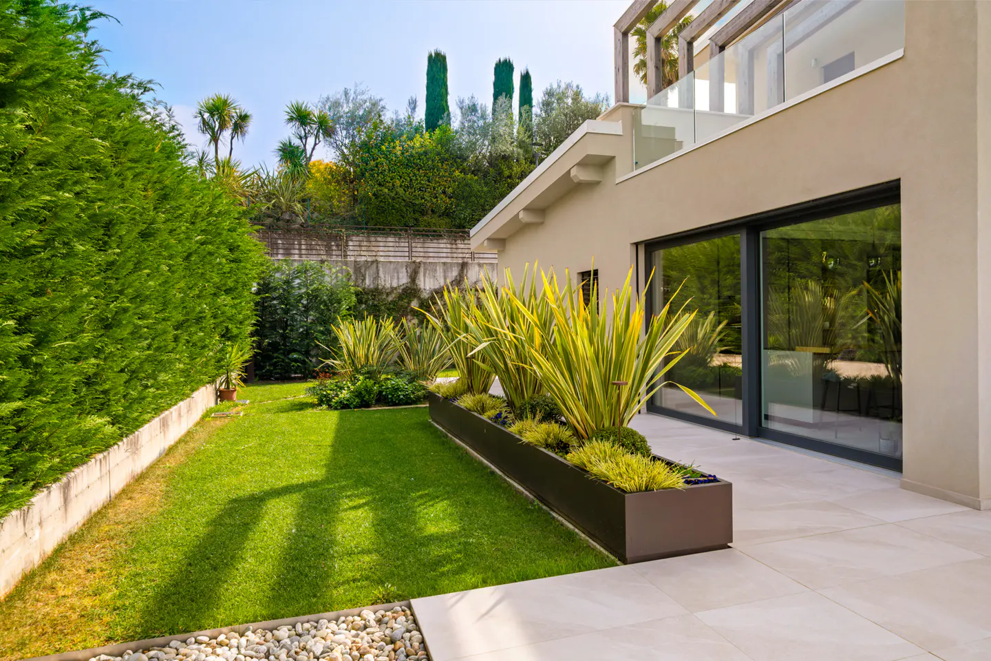 A modern home's backyard with a green lawn, a planter box with yellow plants, and a beige building with sliding glass doors.