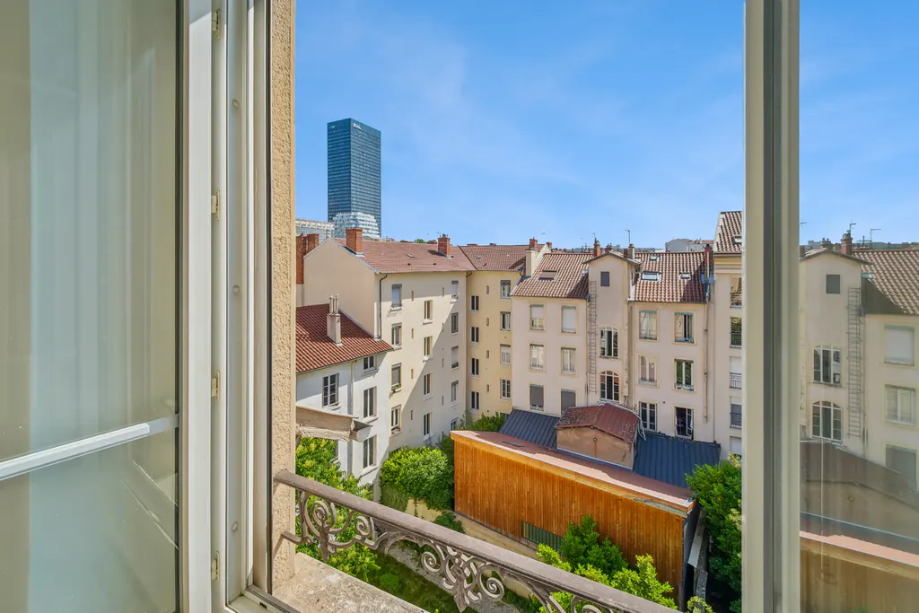 View from a window with an ornate railing, overlooking buildings with red tile roofs and a distant skyscraper under a blue sky.