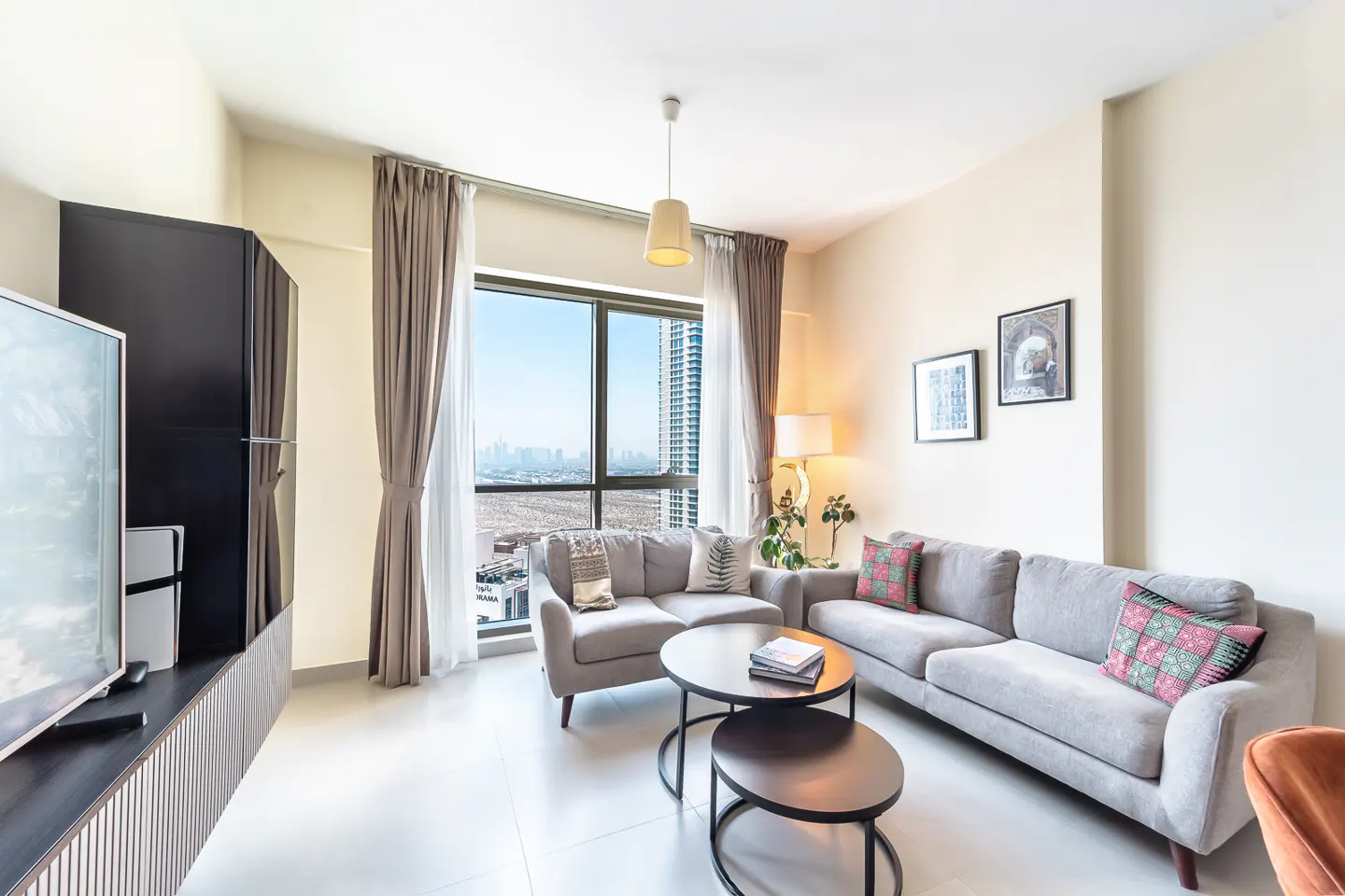 Bright living room with gray sofas, round tables, and a large window showing a city view. A black TV stand is on the left.
