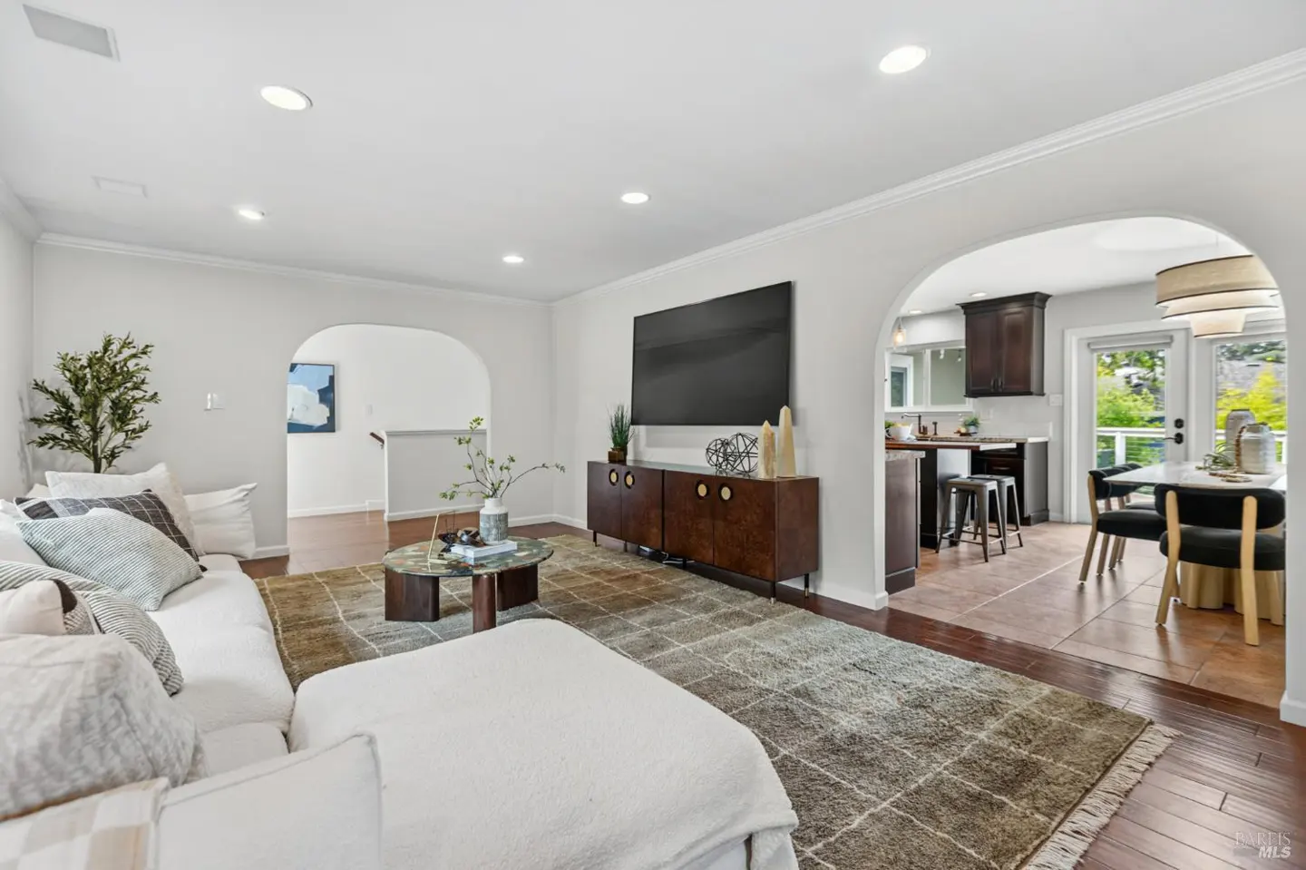 Bright living room with white sofa, patterned rug, and dark wood media console, opening into a kitchen and dining area.