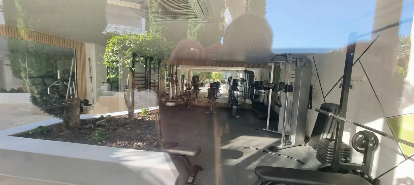 Indoor gym with exercise equipment, including weight machines and stationary bikes. Natural light filters through the windows.