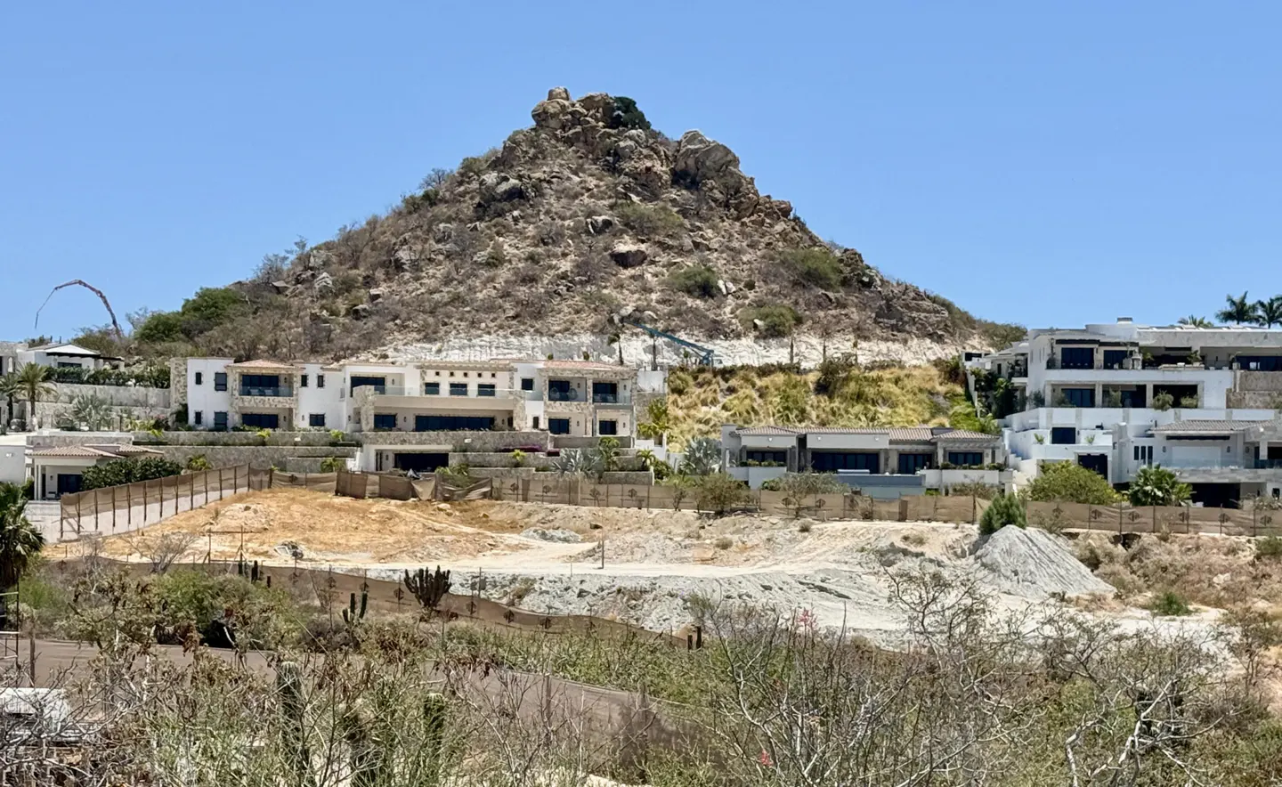 Luxury white villas dot a hillside under a rocky peak against a clear blue sky. Fences and sparse vegetation are in the foreground.