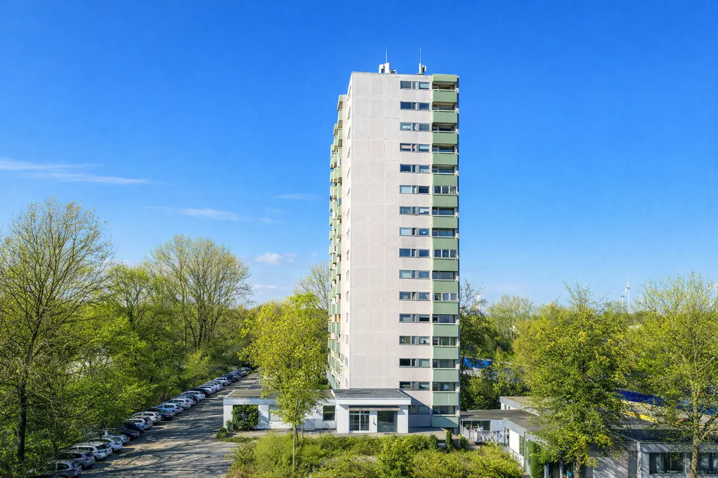 Tall, light gray apartment building with green balconies under a clear blue sky. Trees and a parking lot are in the foreground.