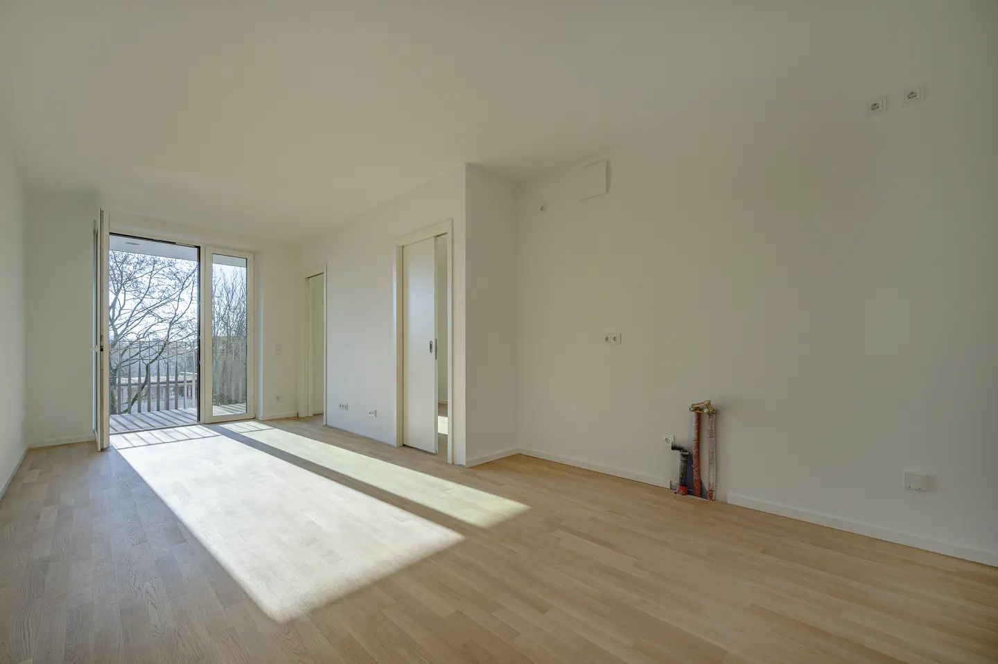 Empty room with light wood floors, white walls, and a glass door leading to a balcony with trees visible outside.