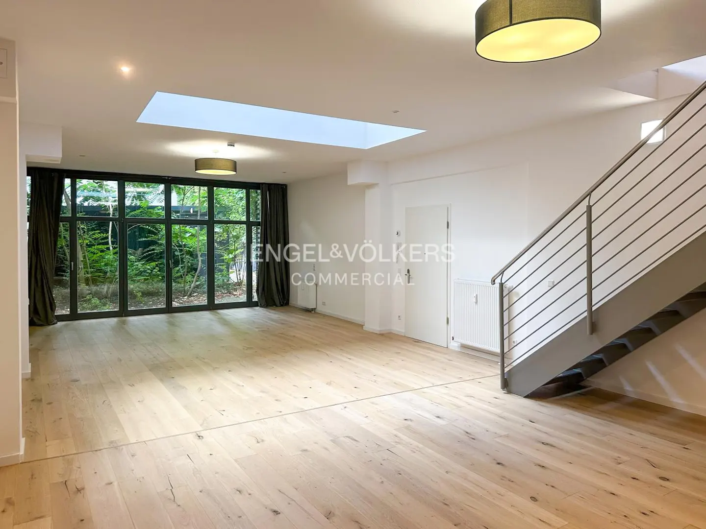 Bright, empty room with wood floors, white walls, and a large window overlooking greenery. Stairs with metal railings are on the right.