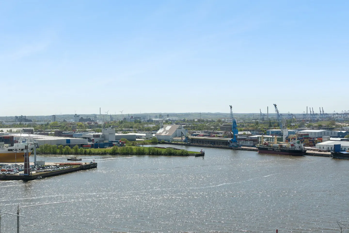 Wide shot of a harbor with cargo ships, cranes, and industrial buildings under a clear blue sky.