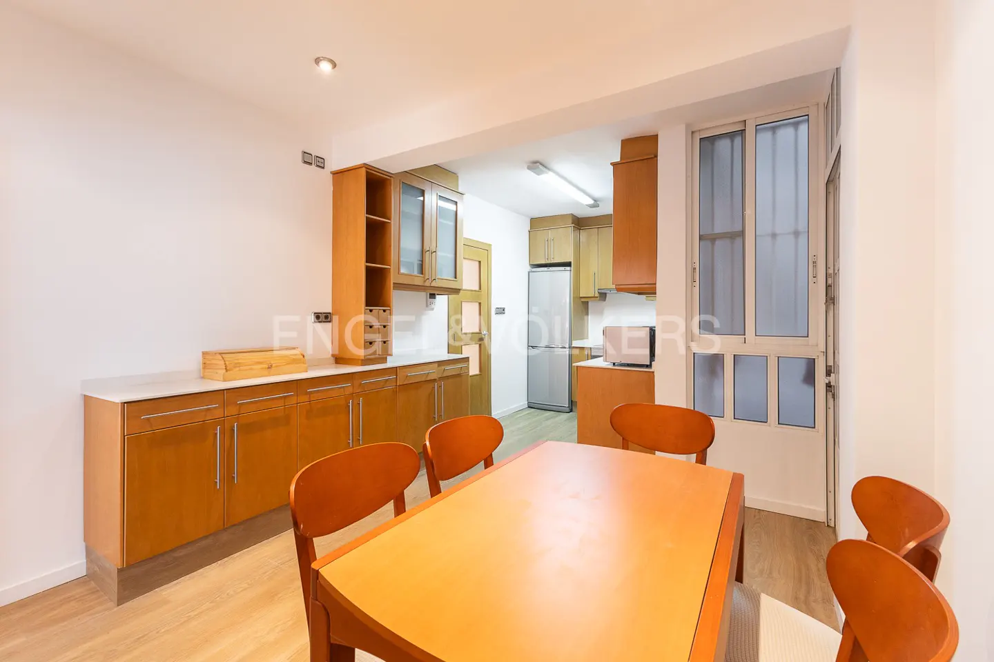 A bright kitchen with wood cabinets, a dining table with orange chairs, and a stainless steel refrigerator.