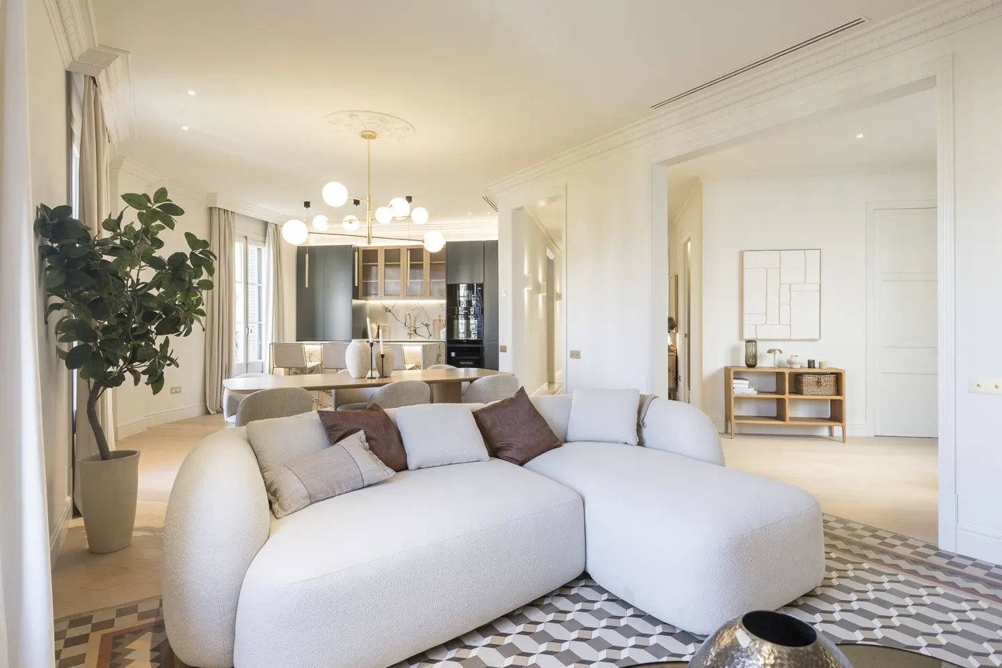 Bright, open-concept living space with a white sectional sofa, patterned rug, and dining area. Kitchen visible in the background.