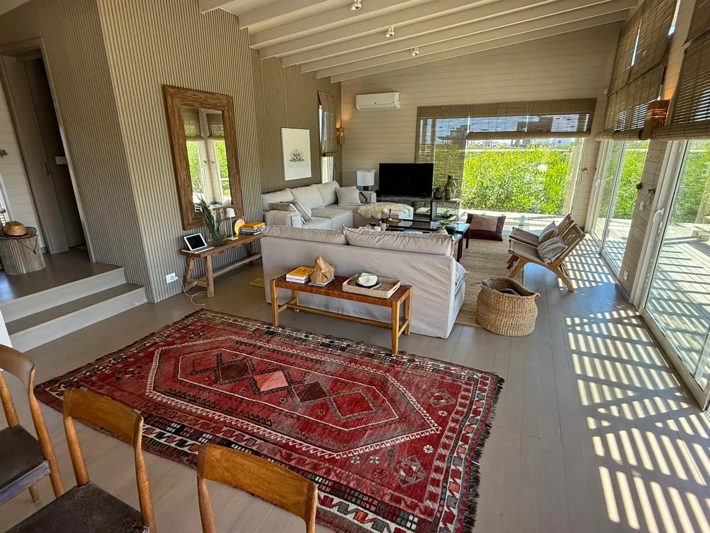 A bright living room with a red rug, a large gray sofa, and floor-to-ceiling windows overlooking green foliage.