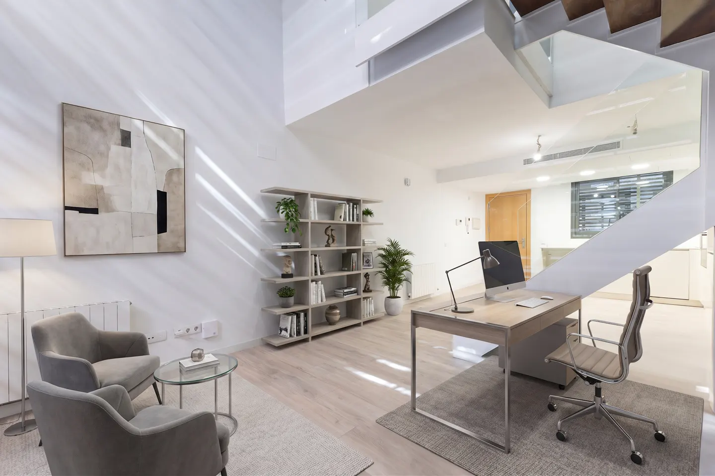 Bright, modern living room with gray chairs, glass table, desk, computer, and bookshelf. White walls and light wood floors. Stairs visible.