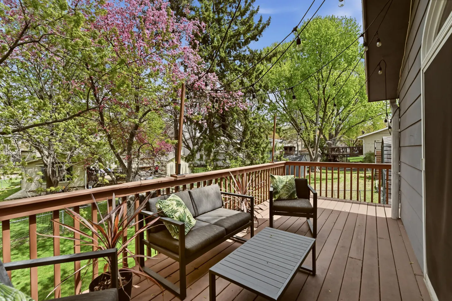 A wooden deck with outdoor furniture, including a sofa, chair, and table, overlooks a lush green backyard with trees and string lights.