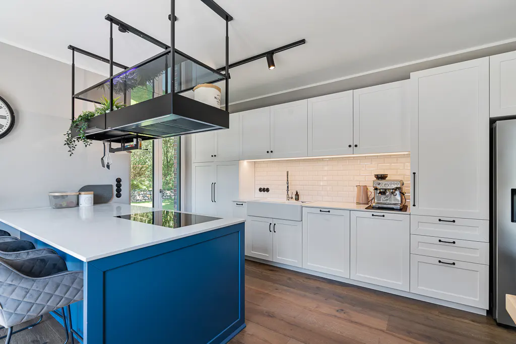 A modern kitchen with white cabinets, a blue island, and hardwood floors. A black range hood hangs above the island.