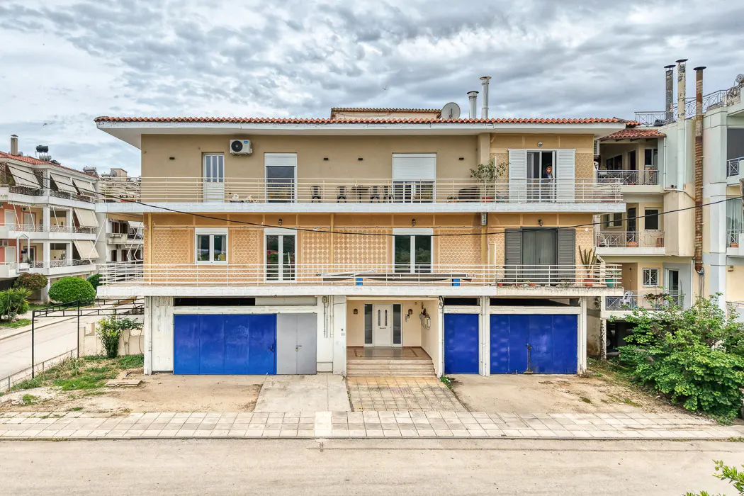 Three-story tan house with blue garage doors and white trim under a cloudy sky. Balconies with white railings are on the upper floors.