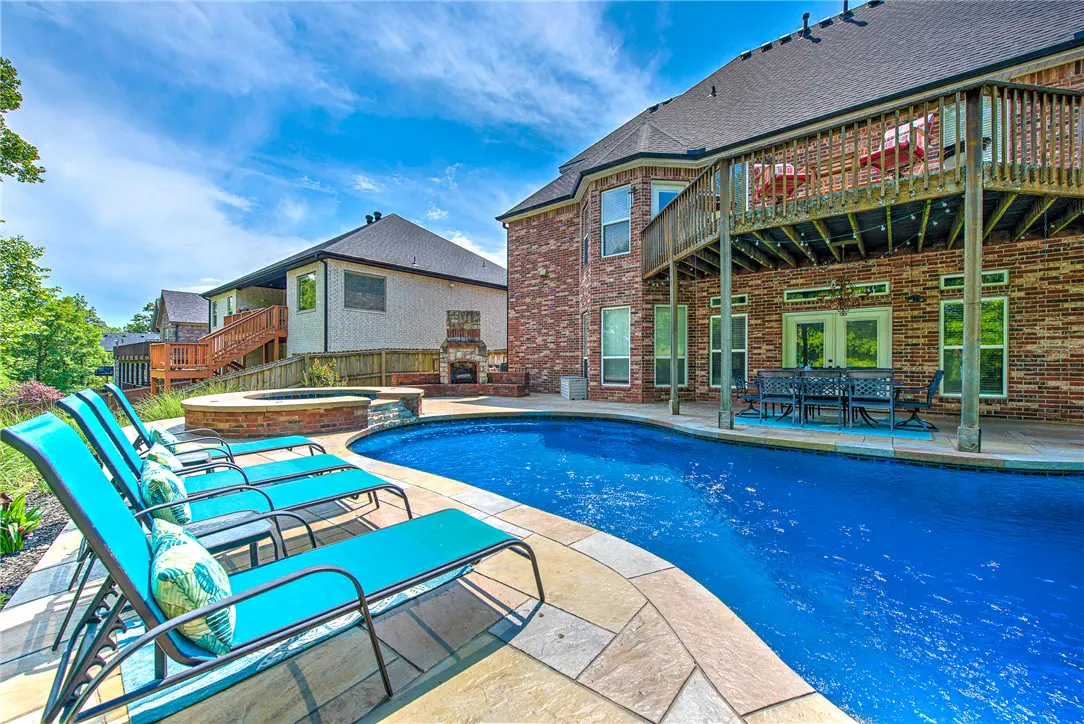 A backyard with a pool, lounge chairs, and a brick house with a deck under a blue sky.