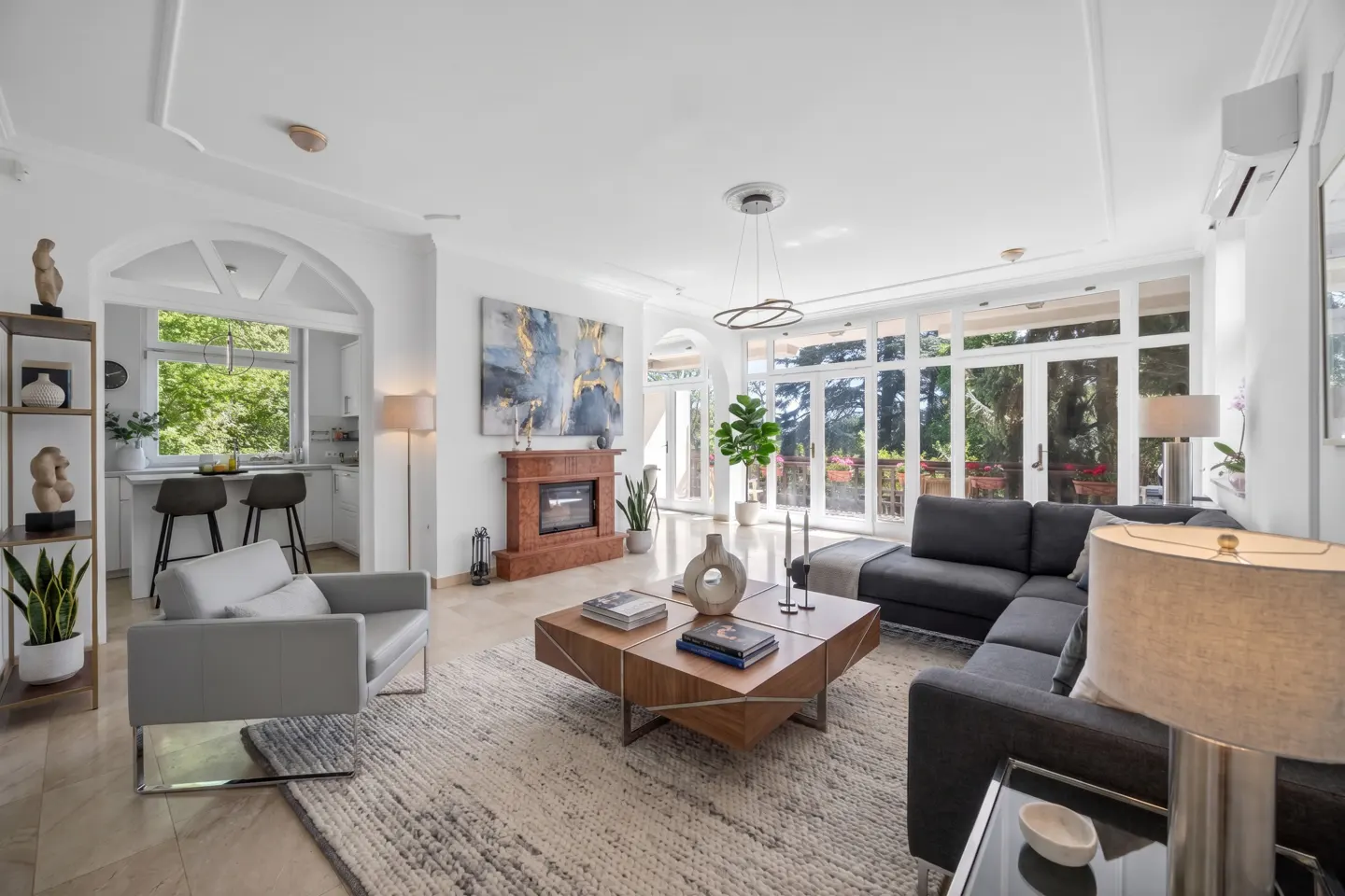 Bright, airy living room with gray sectional, wood coffee table, and fireplace. Large windows overlook a green outdoor space.