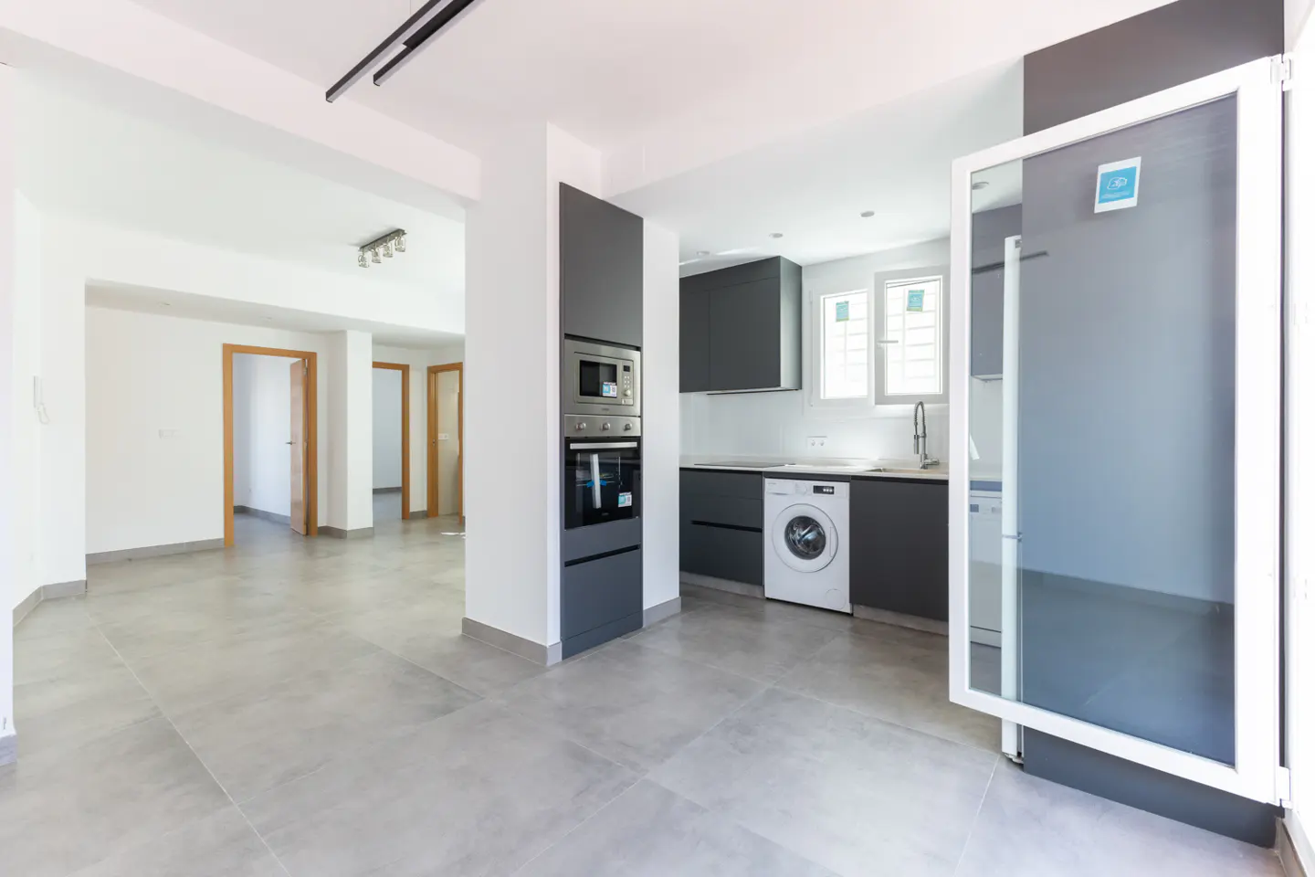 Modern kitchen with dark grey cabinets, built-in oven, and washing machine, opening into a bright, tiled living area.
