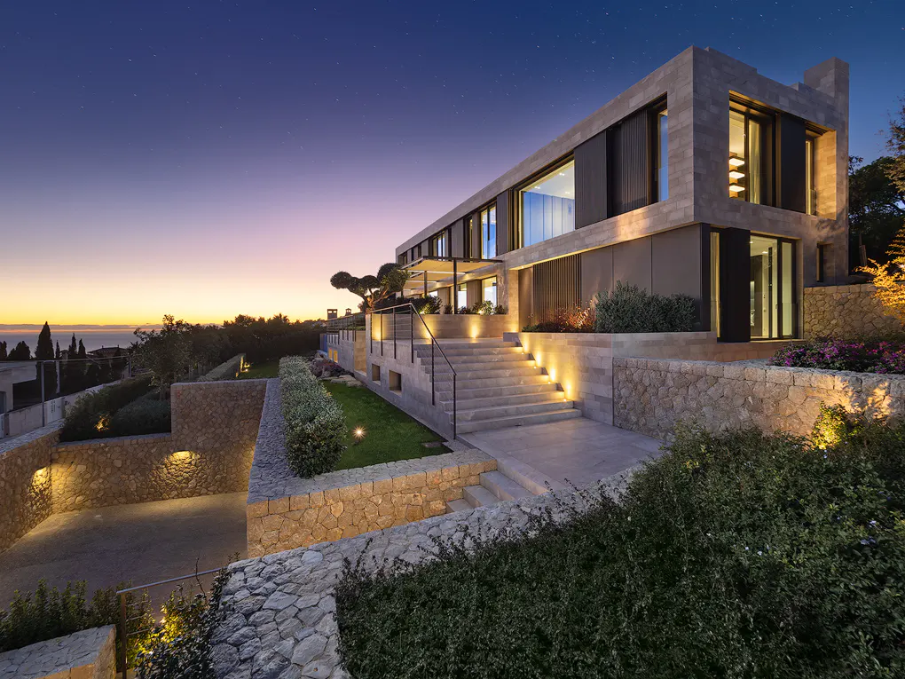 Modern two-story home with stone exterior and large windows at dusk. Steps lead up to the house, with stone walls and greenery in the foreground.
