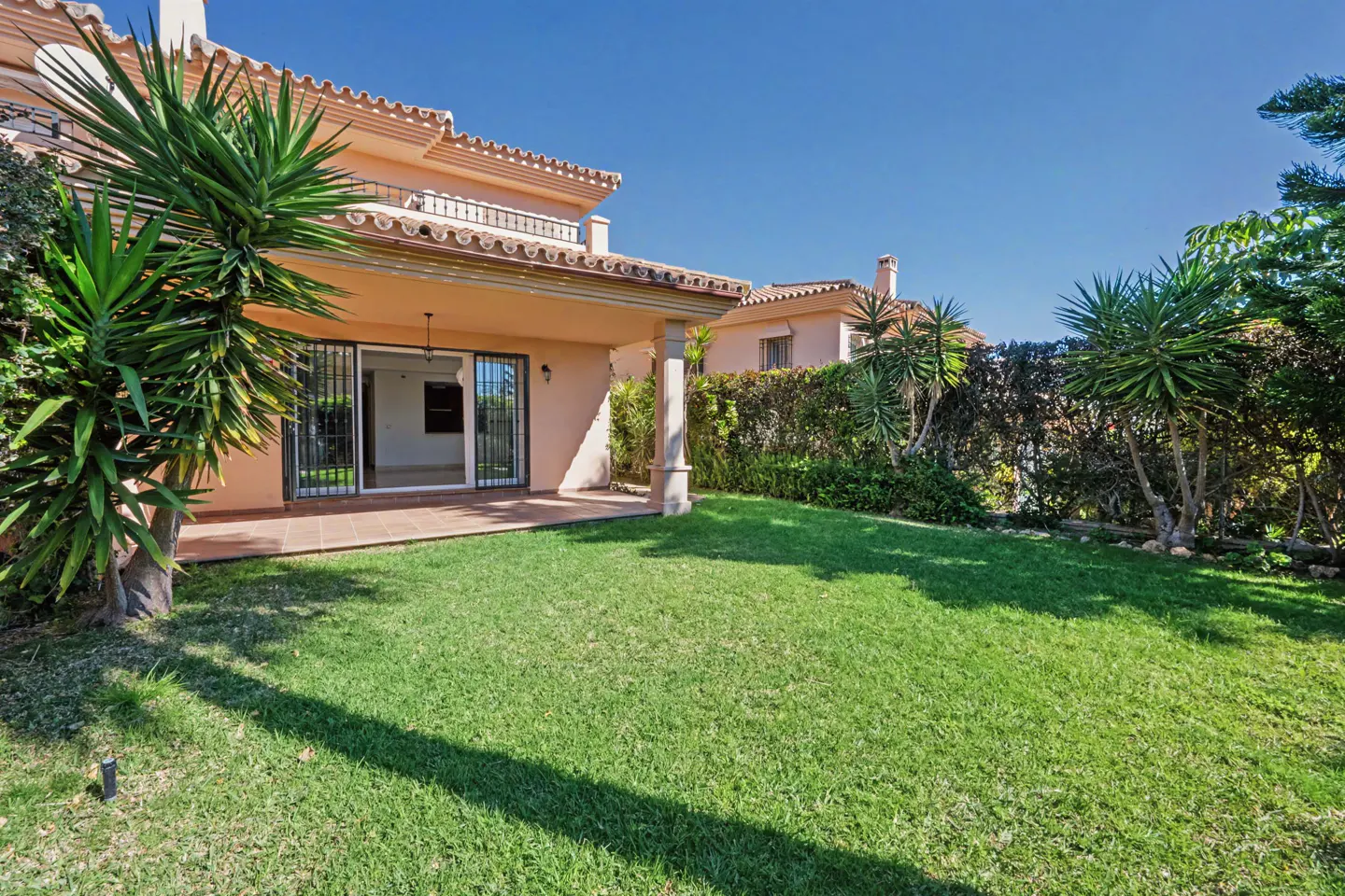 Peach-colored villa with a covered patio, open glass doors, and a lush green lawn under a clear blue sky.